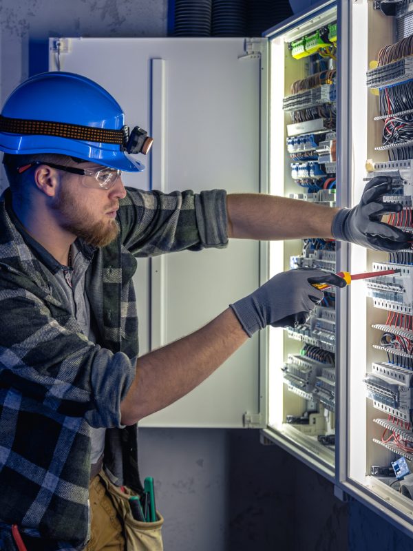 Male electrician working in switchboard with electrical connecting cable. Young adult electrical engineer in special clothes with flashlight on helmet in dark room with emergency lights in background.